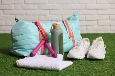 Photo of Stylish sports bag with skipping rope, towel, bottle and sneakers on green grass against white brick wall, closeup