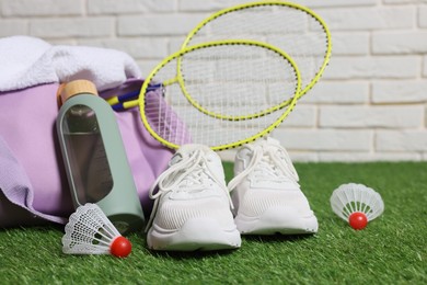 Photo of Stylish sports bag with badminton rackets, shuttlecocks, bottle and sneakers on green grass against white brick wall, closeup