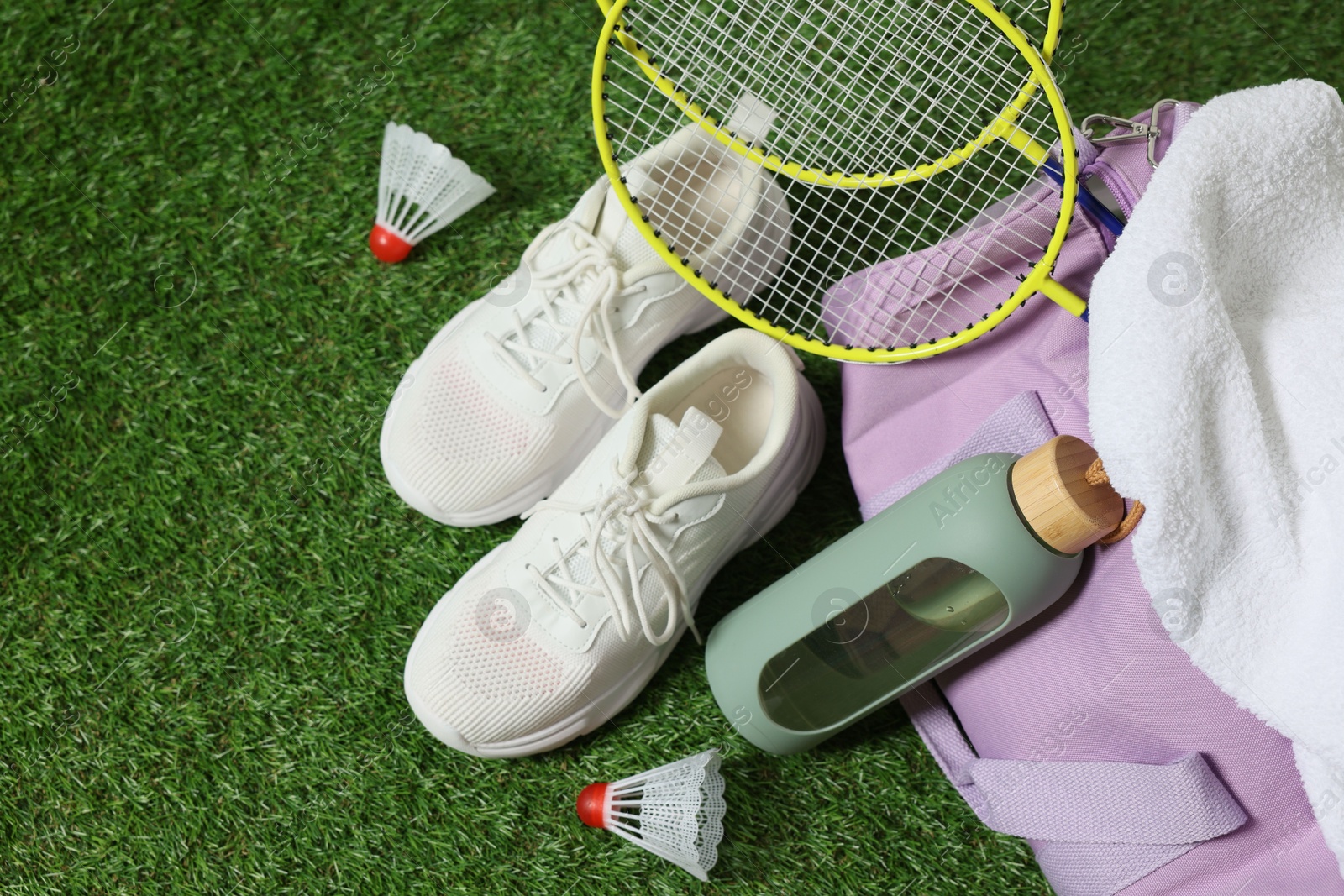Photo of Stylish sports bag with badminton rackets, shuttlecocks, bottle and sneakers on green grass, flat lay