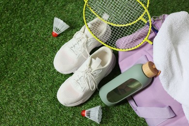 Photo of Stylish sports bag with badminton rackets, shuttlecocks, bottle and sneakers on green grass, flat lay
