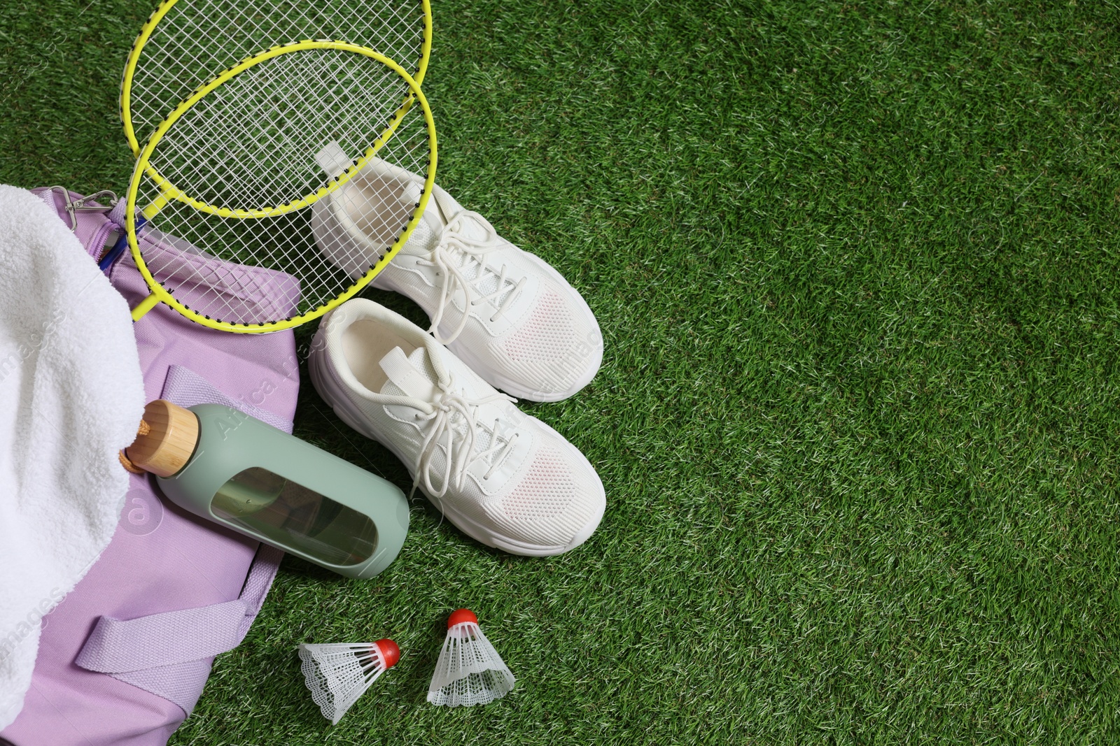Photo of Stylish sports bag with badminton rackets, shuttlecocks, bottle and sneakers on green grass, flat lay. Space for text