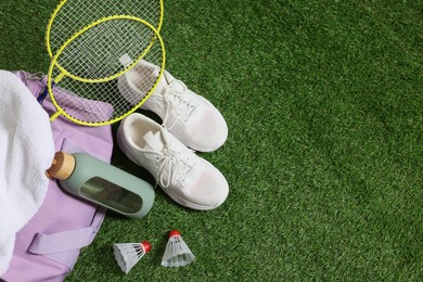 Photo of Stylish sports bag with badminton rackets, shuttlecocks, bottle and sneakers on green grass, flat lay. Space for text