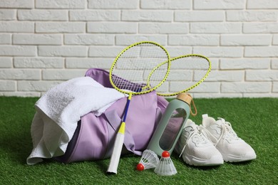 Photo of Stylish sports bag with badminton rackets, shuttlecocks, bottle and sneakers on green grass against white brick wall