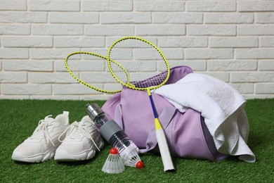 Photo of Stylish sports bag with badminton rackets, shuttlecocks, bottle and sneakers on green grass against white brick wall
