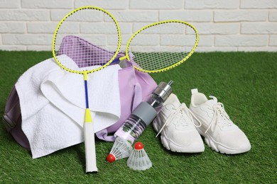 Photo of Stylish sports bag with badminton rackets, shuttlecocks, bottle and sneakers on green grass against white brick wall