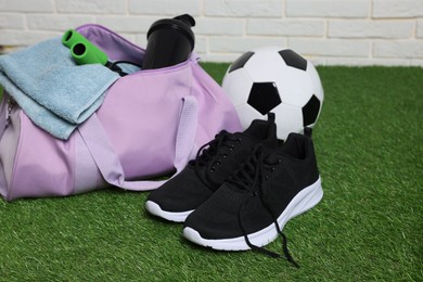Photo of Stylish sports bag, soccer ball and sneakers on green grass against white brick wall, closeup