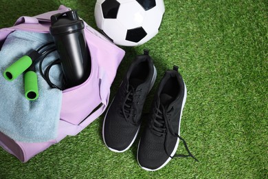 Photo of Stylish sports bag with shaker cup, skipping rope, soccer ball and sneakers on green grass, flat lay