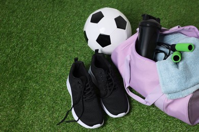 Photo of Stylish sports bag with shaker cup, skipping rope, soccer ball and sneakers on green grass, closeup. Space for text