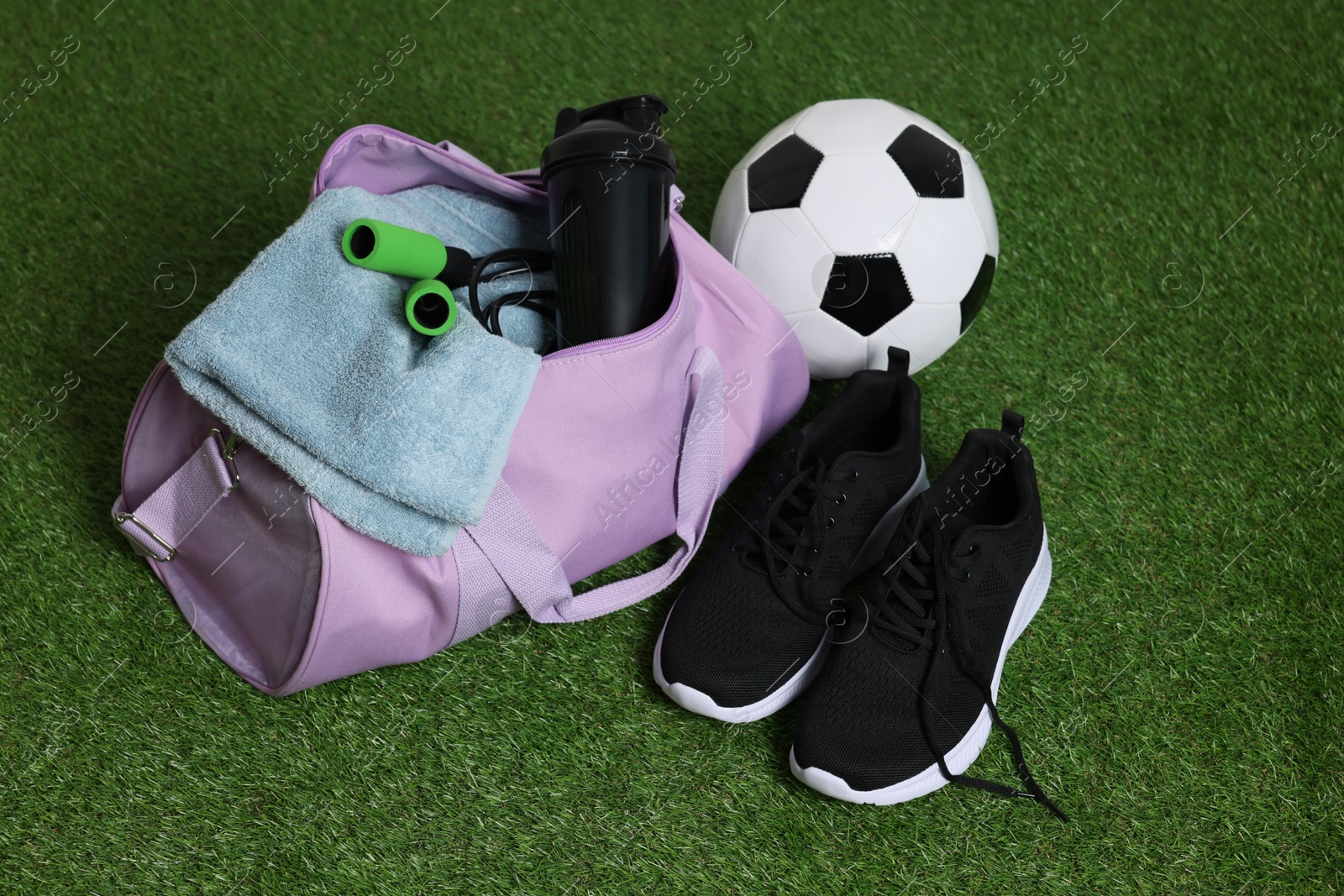 Photo of Stylish sports bag with shaker cup, skipping rope, soccer ball and sneakers on green grass, closeup