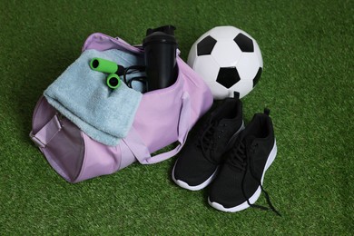 Photo of Stylish sports bag with shaker cup, skipping rope, soccer ball and sneakers on green grass, closeup
