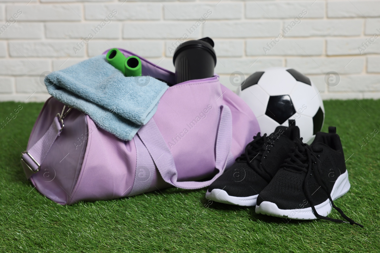 Photo of Stylish sports bag, soccer ball and sneakers on green grass against white brick wall, closeup