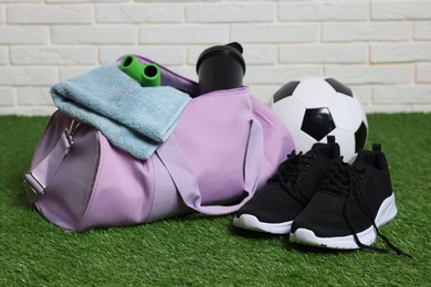 Photo of Stylish sports bag, soccer ball and sneakers on green grass against white brick wall, closeup