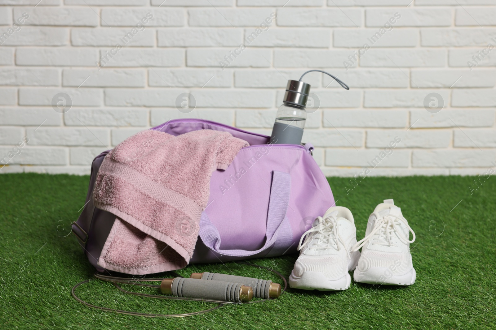 Photo of Stylish sports bag with bottle, towel, skipping rope and sneakers on green grass against white brick wall, closeup