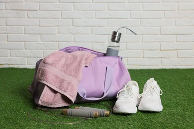 Photo of Stylish sports bag with bottle, towel, skipping rope and sneakers on green grass against white brick wall, closeup