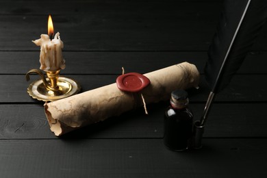 Photo of Old rolled scroll, feather, inkwell and burning candle on black wooden table, closeup