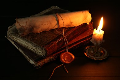 Photo of Old rolled scroll, burning candle and books on black wooden table, closeup