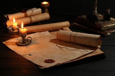 Photo of Many old rolled scrolls, burning candles and books on black wooden table, closeup