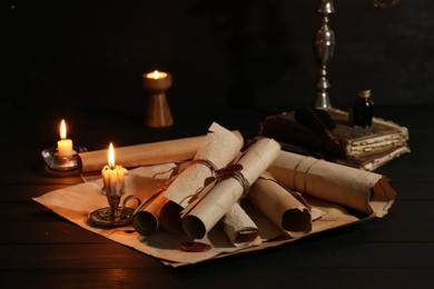 Photo of Many old rolled scrolls, burning candles and books on black wooden table, closeup