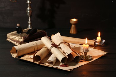 Photo of Many old rolled scrolls, burning candles and books on black wooden table, closeup