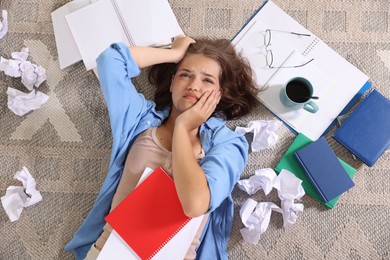 Photo of Tired student among notebooks, crumpled paper and coffee on rug, top view. Preparing for exam
