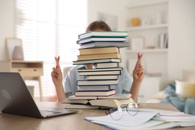 Photo of Student crossing fingers behind stack of books at table indoors. Hope for good exam result