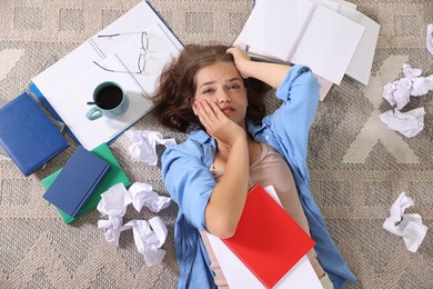 Photo of Tired student among notebooks, crumpled paper and coffee on rug, top view. Preparing for exam
