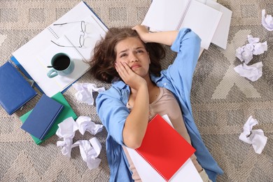 Photo of Tired student among notebooks, crumpled paper and coffee on rug, top view. Preparing for exam
