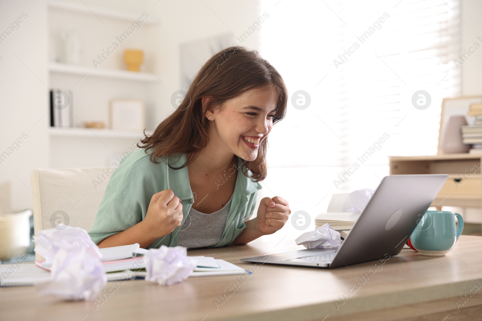 Student feeling happy about her good exam result at table with laptop indoors Photo of Student feeling happy about her good exam result at table with laptop indoors
