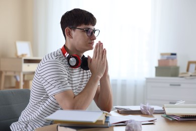 Photo of Worried student with clasped hands praying for good exam result at table indoors