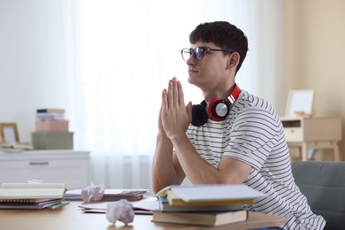 Photo of Worried student with clasped hands praying for good exam result at table indoors