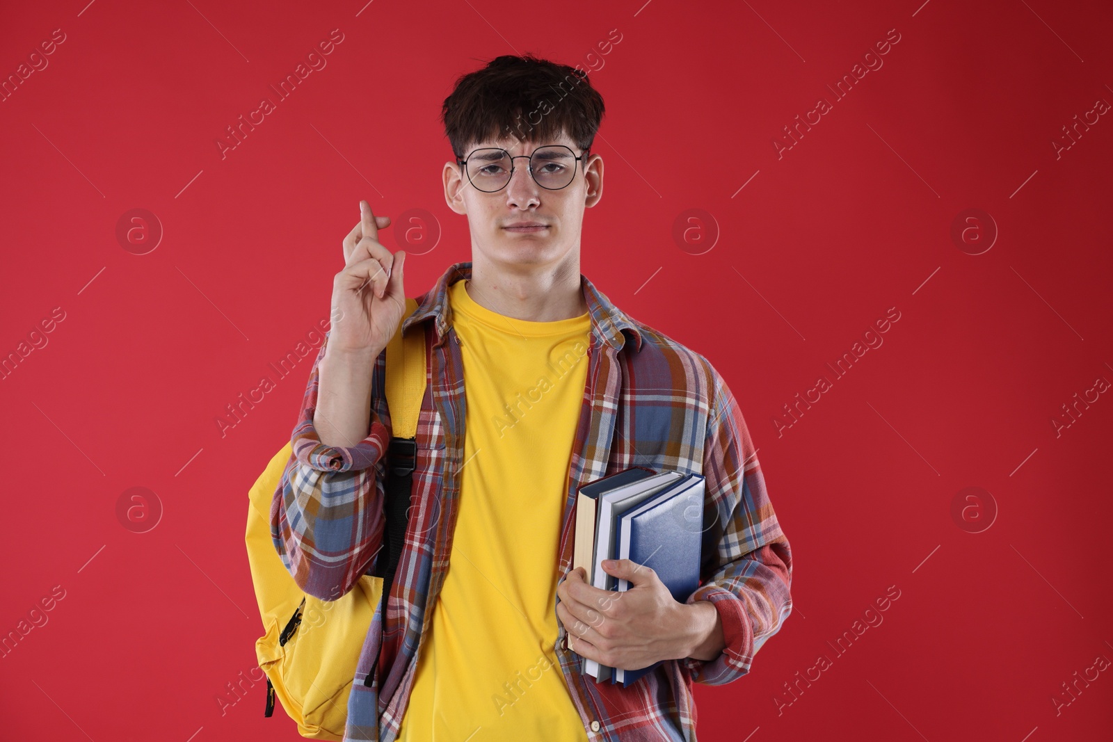 Worried student with books crossing fingers for good exam result on red background Photo of Worried student with books crossing fingers for good exam result on red background