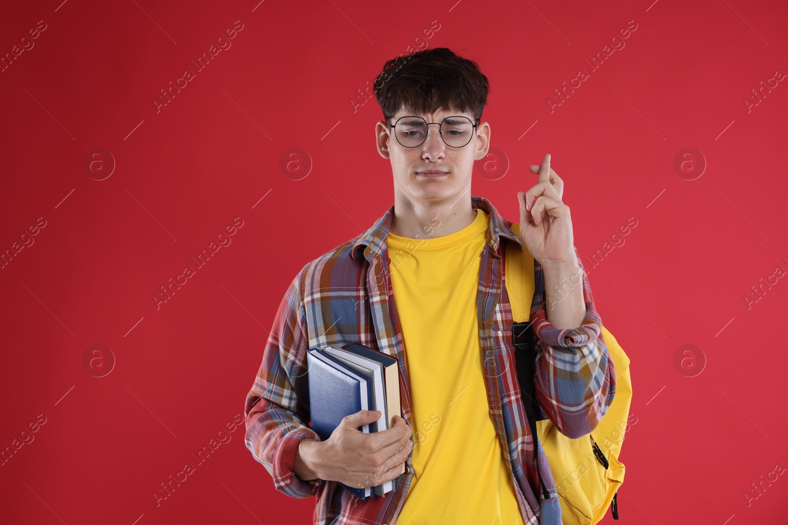 Worried student with books crossing fingers for good exam result on red background Photo of Worried student with books crossing fingers for good exam result on red background