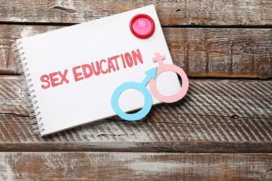 Photo of Notebook with phrase Sex Education, paper female and male gender symbols on wooden table, top view