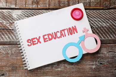 Photo of Notebook with phrase Sex Education, paper female and male gender symbols on wooden table, top view