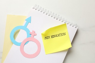 Photo of Sticky note with phrase Sex Education, paper female and male gender symbols in notebook on white table, top view