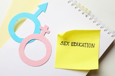 Photo of Sticky note with phrase Sex Education, paper female and male gender symbols in notebook on white table, top view