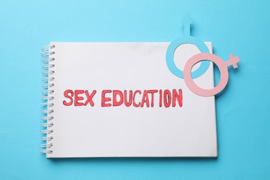 Photo of Notebook with phrase Sex Education, paper female and male gender symbols on light blue background, top view