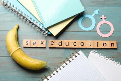 Photo of Phrase Sex Education made of wooden letters, banana with condom, paper female and male gender symbols, notebooks on blue table, flat lay