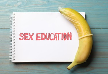 Photo of Phrase Sex Education written in notebook and banana with condom on blue wooden table, top view