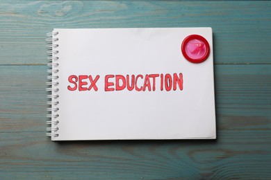Photo of Phrase Sex Education written in notebook and condom on blue wooden table, top view