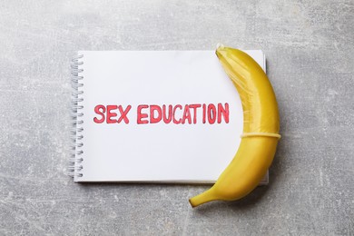 Photo of Phrase Sex Education written in notebook and banana with condom on grey table, top view