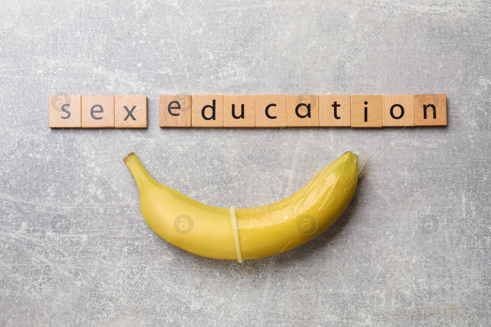 Photo of Phrase Sex Education made of wooden letters and banana with condom on grey table, flat lay
