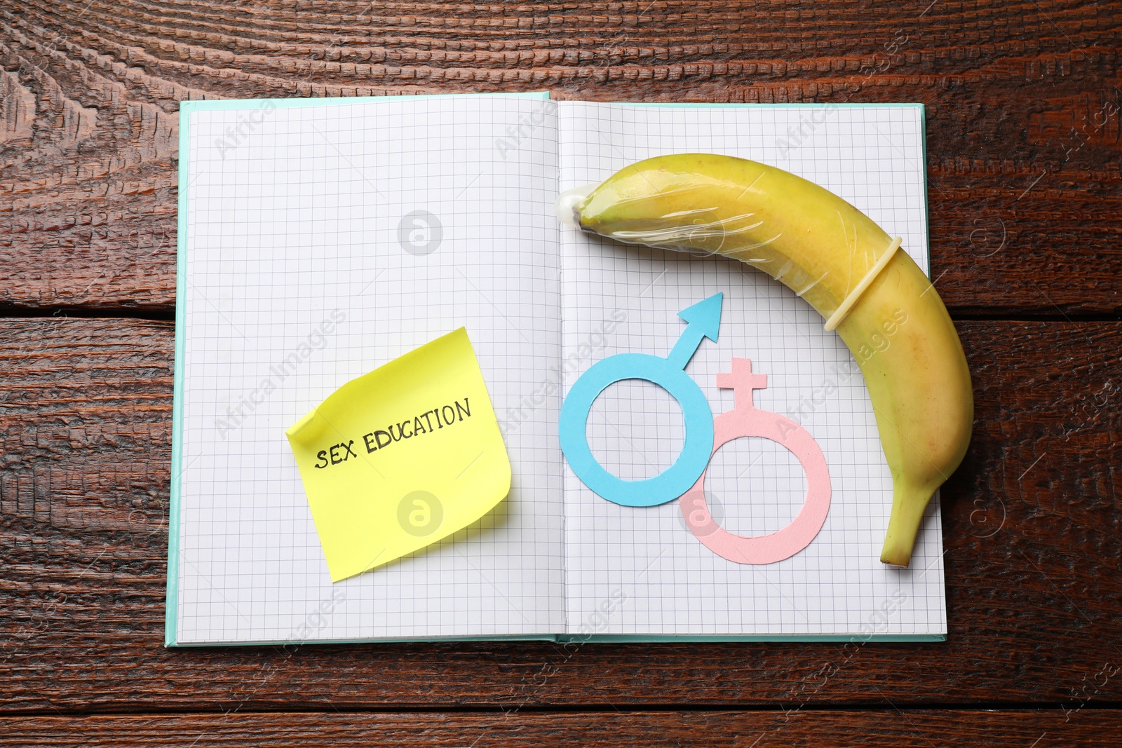 Photo of Sticky note with phrase Sex Education, paper female and male gender symbols, banana with condom in notebook on wooden table, top view