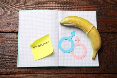 Photo of Sticky note with phrase Sex Education, paper female and male gender symbols, banana with condom in notebook on wooden table, top view
