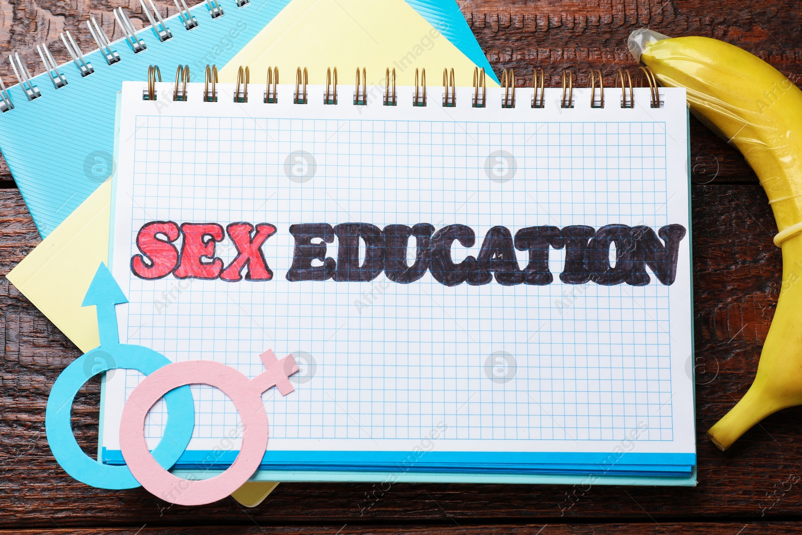 Photo of Notebooks with phrase Sex Education, paper female and male gender symbols, banana with condom on wooden table, flat lay