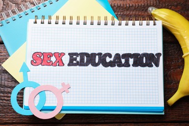 Photo of Notebooks with phrase Sex Education, paper female and male gender symbols, banana with condom on wooden table, flat lay