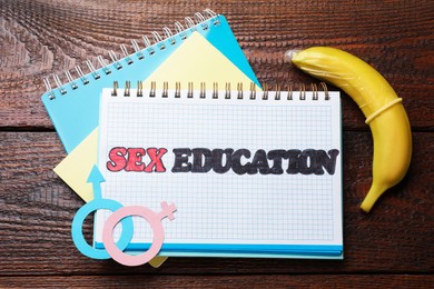 Photo of Notebooks with phrase Sex Education, paper female and male gender symbols, banana with condom on wooden table, flat lay