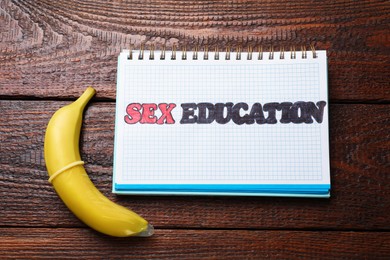 Photo of Phrase Sex Education written in notebook and banana with condom on wooden table, top view