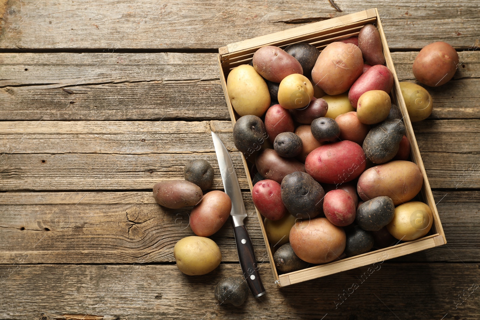 Photo of Different types of potatoes in crate and knife on wooden table, flat lay. Space for text