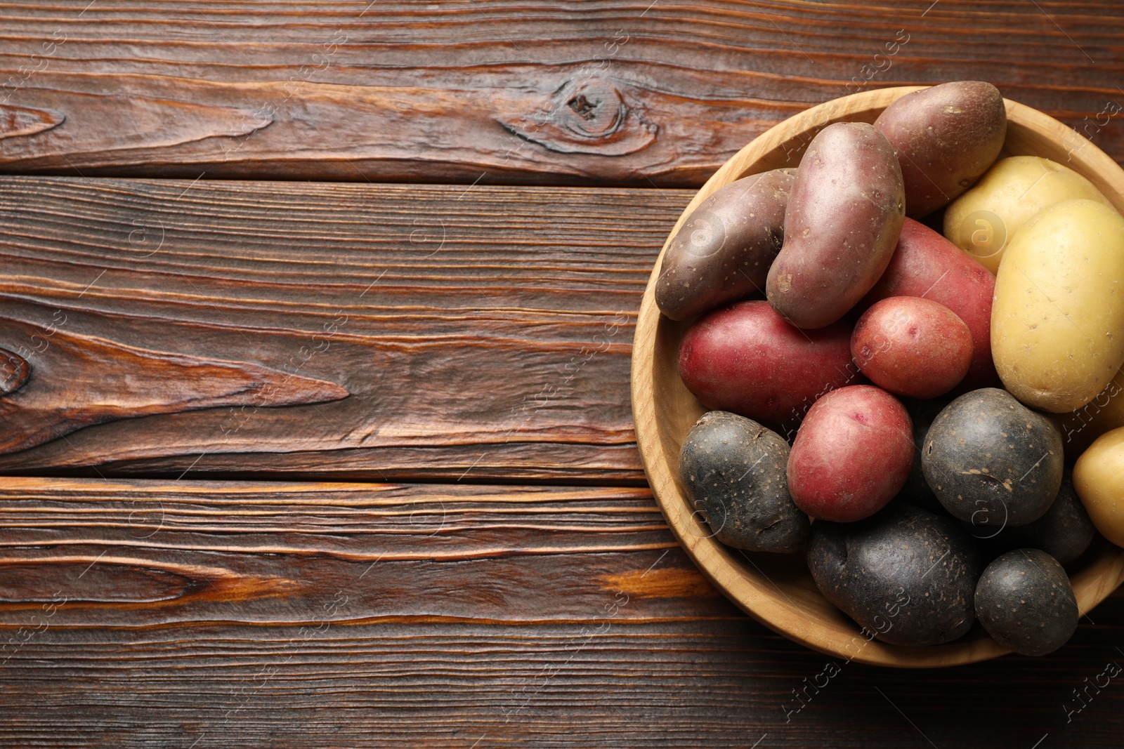 Photo of Different types of potatoes in bowl on wooden table, top view. Space for text